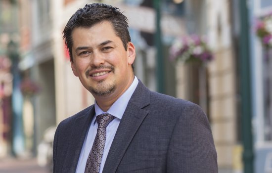 A man stands in front of a bright city street wearing a gray suit, blue shirt and gray/purple tie. He has a goatee and dark brown/black hair.
