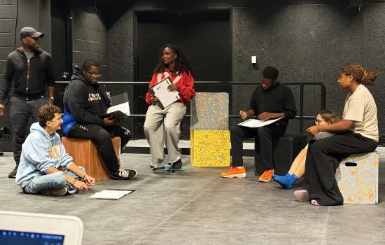 a group of theatre students and faculty, one standing to the left, the rest seated on the floor or boxes, look over scripts and notes in a flat theatre space with dark or black walls behind them.