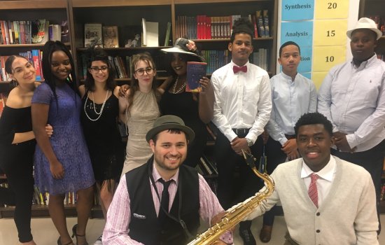 A group of people in 1920s attire pose for a photo. One is holding a saxophone and another is holding The Great Gatsby book.