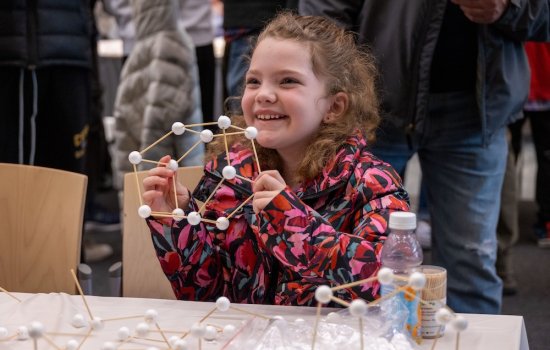 A young girl in a colorful coat grins as she holds up a science craft constructed with sticks and balls.
