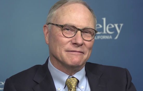 A man with balding hair and glasses, wearing a dark suit and yellow/gold checkered tie sits  for a portrait with a logo of the University of California, Berkeley behind him.
