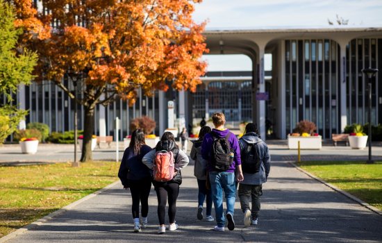 Five students wearing backpacks walk away from the camera down a paved pathway toward UAlbany's academic buildings on a sunny fall day.