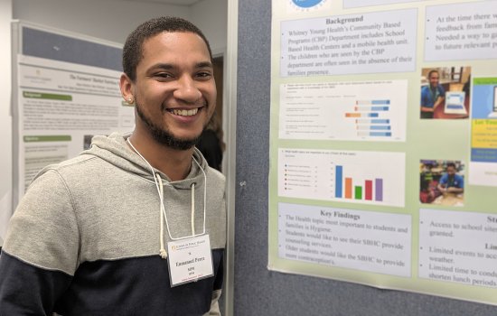 A student stands in front of his poster, which includes small text and lots of colorful graphs and charts. The student is smiling.