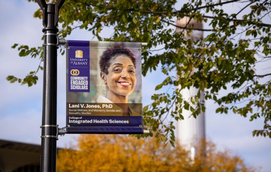 A banner hangs from a light post on campus on a sunny fall day, with the carillon in the background. The banner includes the University at Albany logo and the Community Engaged Scholars logo, as well as a the words, "Lani V. Jones, PhD, Social Welfare and Women's Gender and Sexuality Studies, College of Integrated Health Sciences" and a photo of Lani V. Jones.