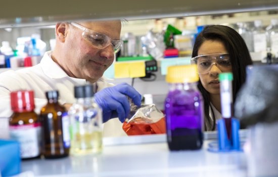 A photo of several glass bottles filled with colorful liquids with two scientists behind them in lab gear.
