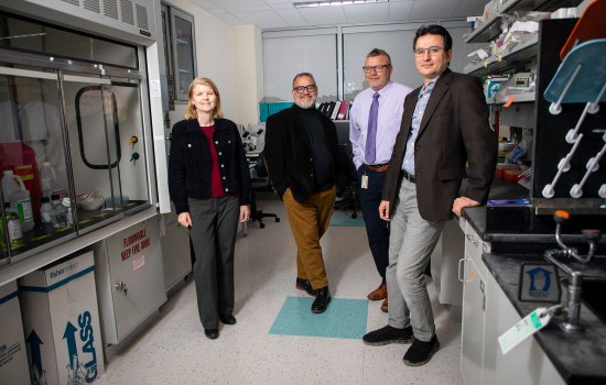 The four featured researchers stand in Melinda Larsen's lab in the RNA Institute. The room itself is dimly lit, with the camera flash adding light. From left to right: Melinda Larsen (woman with blond hair wearing a black jacket with gray pants), Scott Tenenbaum (man with black glasses wearing black jacket with khaki pants), Andre Melendez (man wearing black rimmed glasses, pale purple dress shirt and tie and black slacks, and Mehmet Yigit (man wearing wire rimmed glasses, brown jacket and grey pants).