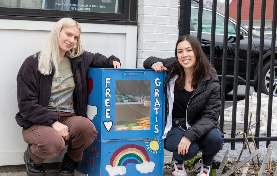 Two smiling young women crouch on a sidewalk next to a blue metal box containing period products. A rainbow and the words “Free” and “Gratis” are painted on the front of the box. A glass window displays period products inside.