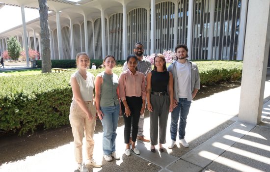 Six people, including four women and two men, stand together, smiling for a group portrait. They are standing outside, sheltered by the roof of UAlbany's academic podium. A row of evergreen bushes and academic buildings can be seen in the background.