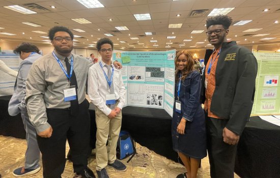 Four students — three boys and a girl wearing name tags and dress clothes — stand next to a poster board depicting air pollution research in a hotel conference room.