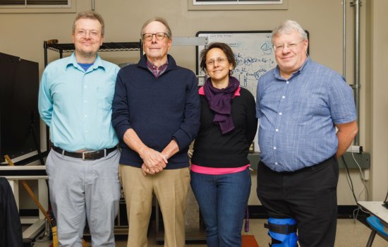 three men and a woman stand in a row facing the camera in front of a whiteboard scrawled with figures and shapes