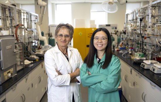 A older woman with short light brown hair and glasses in a white lab coat stands with a younger woman with long dark hair, glasses and a green lab coat in a chemistry laboratory with tools, beakers and chemicals.