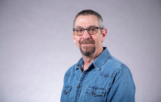 A man with short gray hair, a goatee and glasses wears a denim jacket and poses for a portrait against a gray backdrop.