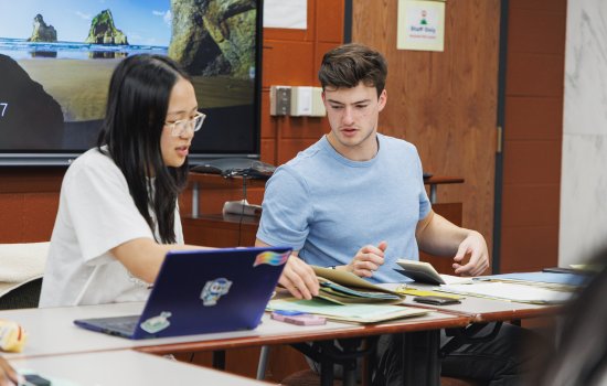 A young woman with dark hair, glasses and a white T-shirt sits next to a young man with brown hair and a blue t-shirt at a table as they go through folders of paper.