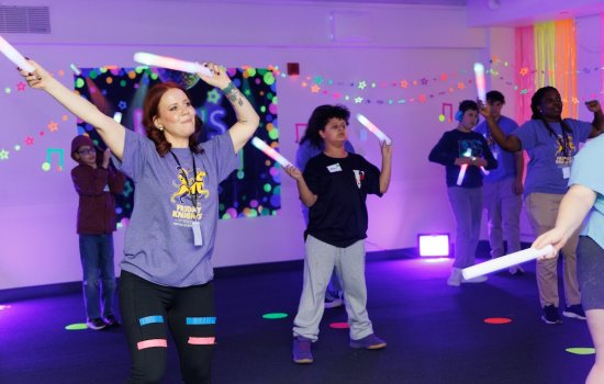 Children and adults wave glow sticks in a room that is lit purple and decorated with glow-in-the-dark stars and tape.