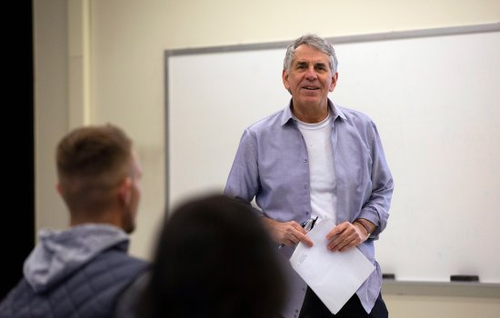 A gray-haired professor stands in front of a class holding his glasses and some papers.