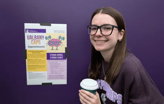 Izzy D'Ambro stands next to a small poster display of her social media messages for making an appointment at UAlbany's Counseling Center. She holds a cup of coffee.