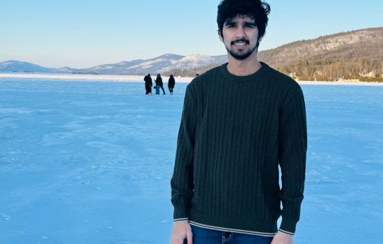 A man in a dark sweater stands on a frozen lake under a clear blue sky. Mountains line the horizon, with four distant people walking behind.