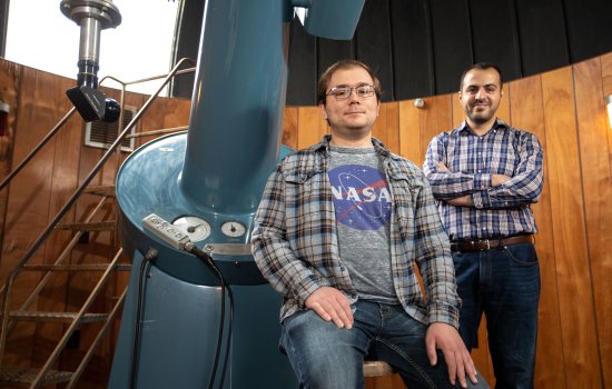 UAlbany PhD student John Bradburn sits by a large telescope in an observatory as Assistant Professor Mustafa Aksoy stands in the background