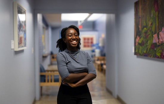 A young woman smiles broadly, arms crossed, standing in a lavender-painted hallway lined with colorful art pieces.