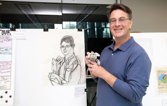 A man wearing glasses and a blue shirt holding a small stuffed mouse toy stands in front of a portrait of himself holding a mouse sketched on paper.