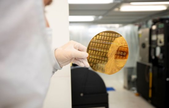 A gloved hand holds a gold-colored silicon wafer reflecting the image of a person in a cleanroom "bunny" suit.