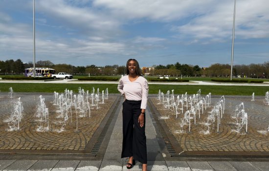 A person standing confidently near a fountain with water jets in a park, under a partly cloudy sky. Green grass and trees are visible in the background.