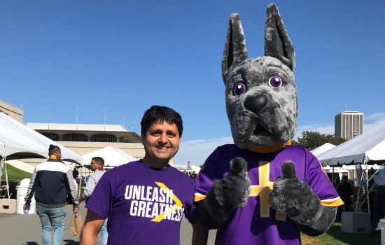 Khem Sedhai, wearing a purple t-shirt with the UAlbany slogan, "Unleash Greatness," smiles and poses for a photo with the UAlbany mascot, Damien the Great Dane.