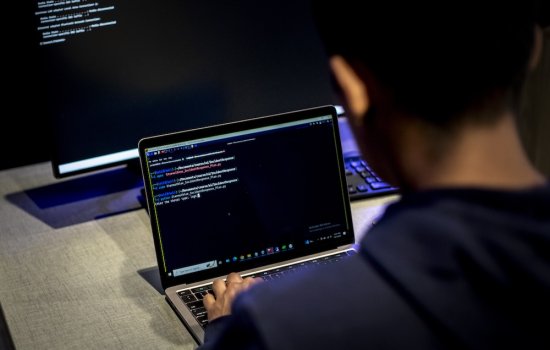 A man sits at a desk with a desktop computer and types code on a laptop.