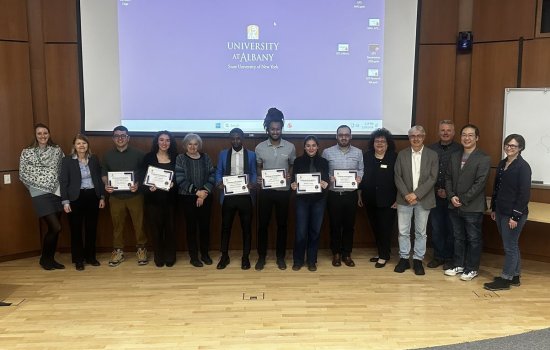 Fourteen people pose for a group portrait in front of a pull-down projection screen displaying the UAlbany logo. All are smiling and six are holding white paper certificates.