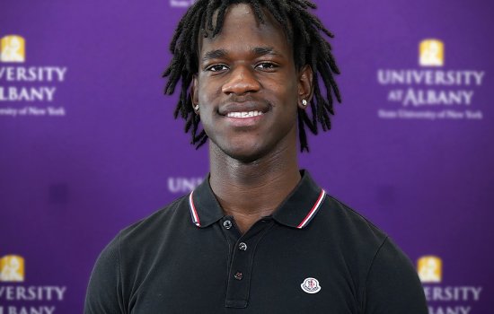 An African Americna young man wears a black collared shirt while standing against a purple backdrop with University at Albany written on it.