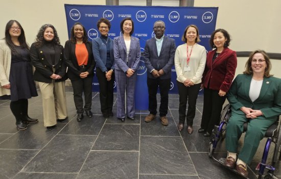 A group of University administrators, faculty and staff pose in front of a blue banner with the words SUNY and State University of New York repeated in white letters.