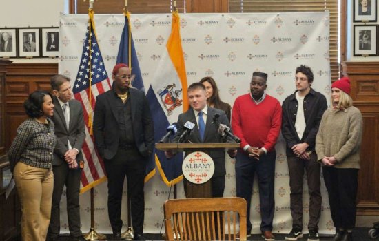 A young man in a suit speaks at a lecturn with a round plaque reading ALBANY on it, as 7 others gather around watching him