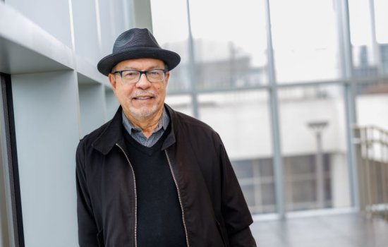 A portrait of a man with eyeglasses in a dark gray hat, black sweater and black jacket leaning against a wall in atrium filled with natural light.