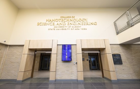 A sign for the College of Nanotechnology, Science, and Engineering hangs above two doorways in a building with light tan and brick walls.