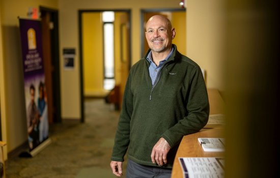 A man wearing a green quarter-zip sweater smiles for a portrait in a warmly lit hall.