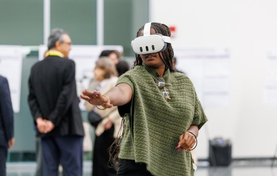 A woman with long braids and a green sweater wears a virtual reality headset and gestures in front of her.