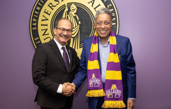 A smiling man in a blue blazer, gold and purple UAlbany scarf shakes hands with a smiling man in a black suit and purple tie.