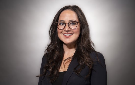 A woman with long dark hair and glasses in a black blazer smiles for a portrait against a gray backdrop.