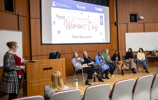 Seven women are seated on a stage as another woman asks questions from a lectern. A screen behind them says "International Women's Day Panel Discussion.""