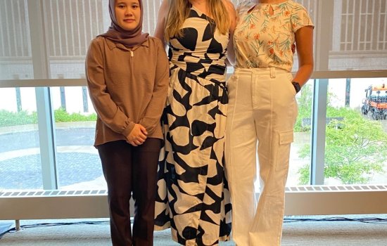 Three women stand smiling indoors by large windows. The first wears a hijab and brown outfit, the second a black and white dress, and the third a floral top and white pants. The atmosphere is friendly and bright.