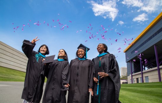 Four graduates in black gowns and caps celebrate outdoors under a blue sky. Purple confetti falls as they smile, exuding joy and accomplishment.