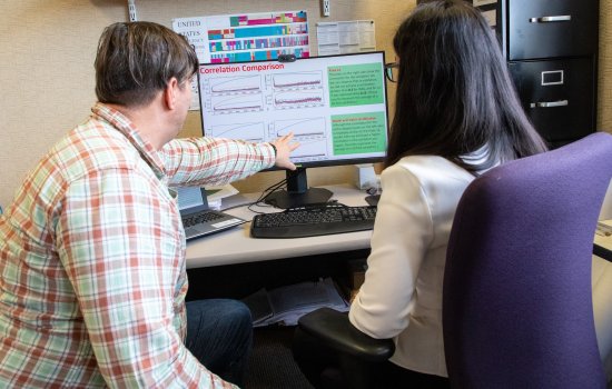 Professor seated next to student points to computer screen in front of them on a desk. 