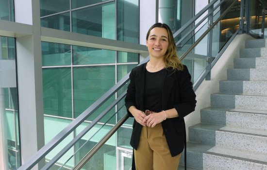 A young woman with brown-blond ombre hair, wearing a black top, black blazer and khaki slacks, smiles for a portrait in a stairwell with glass walls and gentle natural light.