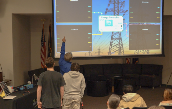 A man at the front of an auditorium points to a screen demonstrating the energy input and output levels of a solar array simulator.