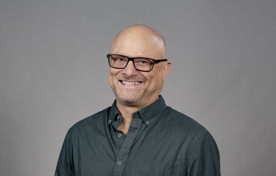 A man wearing a slate button-down shirt and glasses with black rectangular frames smiles for a portrait against a gray backdrop.