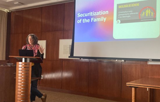 An image of Dr. Annie Wilkinson standing in front of a podium, a drop-down screen behind her with a "Securization of the Family" slide.