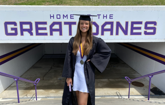 A young woman with long brown hair in a white dress and black graduation cap and gown poses in front of a sign that says "Home of the Great Danes"