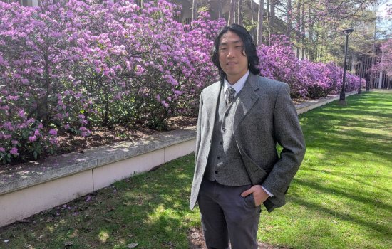 A young man with dark hair wearing a gray blazer and gray khaki pants poses for a portrait in front of a magnificent hedge of purple azaleas in full bloom on a sunny day.
