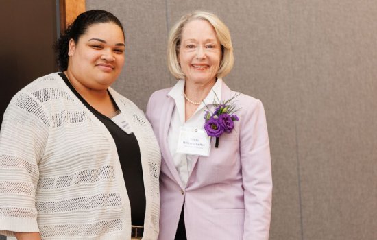 Two smiling women stand together wearing nametags. The older woman has flowers in her lapel.