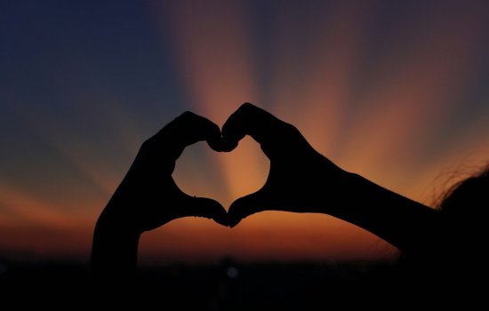 Two hands form a heart shape in front of a sunset, creating a silhouette.